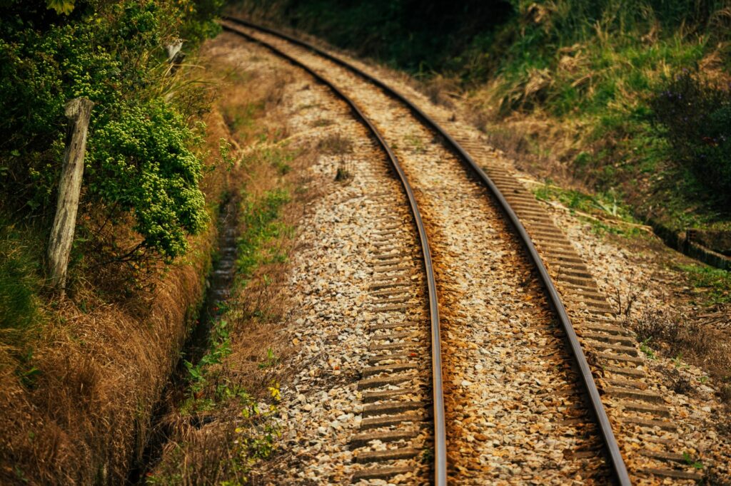 Trilhos de trem em uma curva em meio a natureza, com dormentes de madeira, trilho de aço e vegetação nas laterais, vistos em perspectiva.
