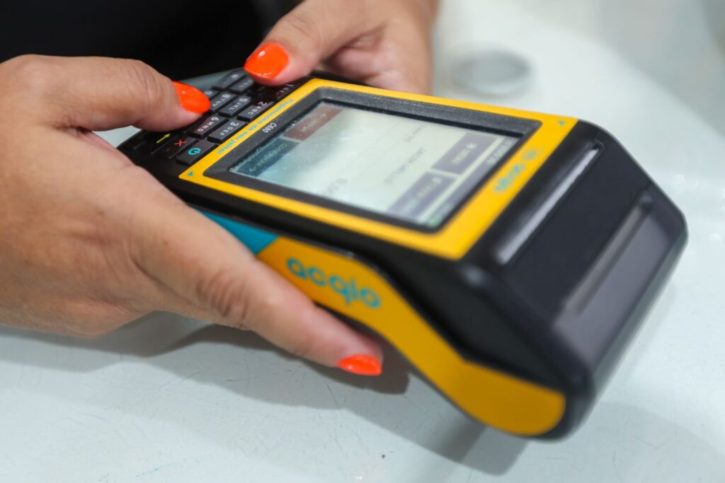 Pessoa utilizando um leitor de código de barras portátil amarelo e preto, com teclado e tela sensível ao toque, enquanto pressiona botões com as mãos, em ambiente de atendimento.