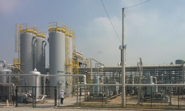 Complexo industrial com tanques de armazenamento e tubulações metálicas amarelas, tubulações e equipamentos de processo ao ar livre, em uma planta com estruturas e cercas.
