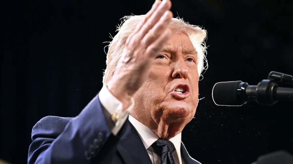 Donald Trump aparece em um palco durante um discurso, com a mão levantada em gesto de cumprimento ou ênfase, ao lado de um microfone e com expressão séria.