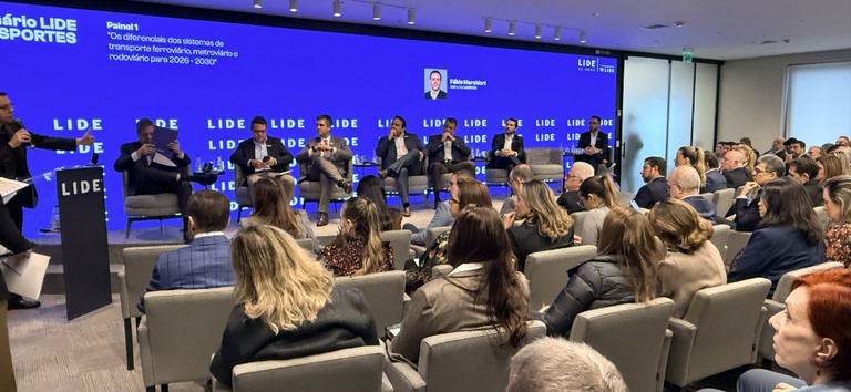 Evento corporativo com palestrantes em palco e público sentado em auditório, diante de um painel azul com a marca LIDE e telão de apresentações