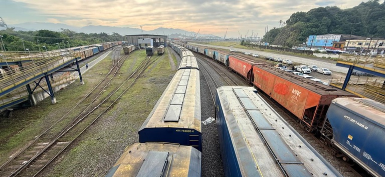 Vista aérea de um pátio ferroviário com trilhos paralelos, vagões de carga e um trem estacionado, ao fundo morros e estrutura industrial na paisagem.