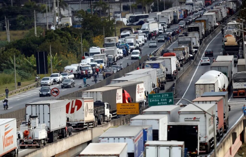Trânsito intenso em rodovia com grande fila de caminhões e veículos de carga, carros parados em ambas as faixas e diversos veículos pesados ao longo da pista, em dia movimentado.