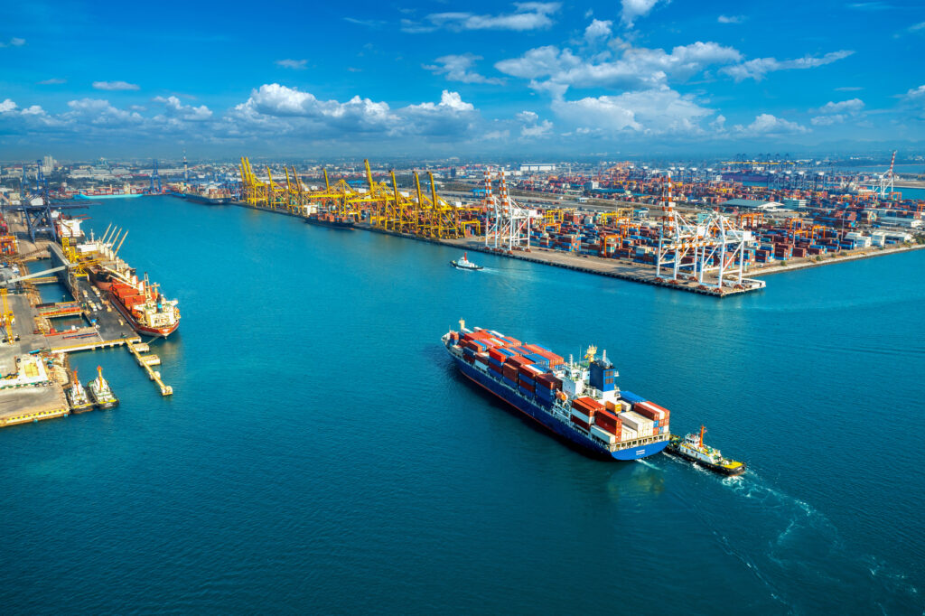 Vista aérea de um porto comercial com contêineres e guindastes amarelos sobre águas azul-esverdeadas, com um navio cargueiro navegando ao centro da imagem.