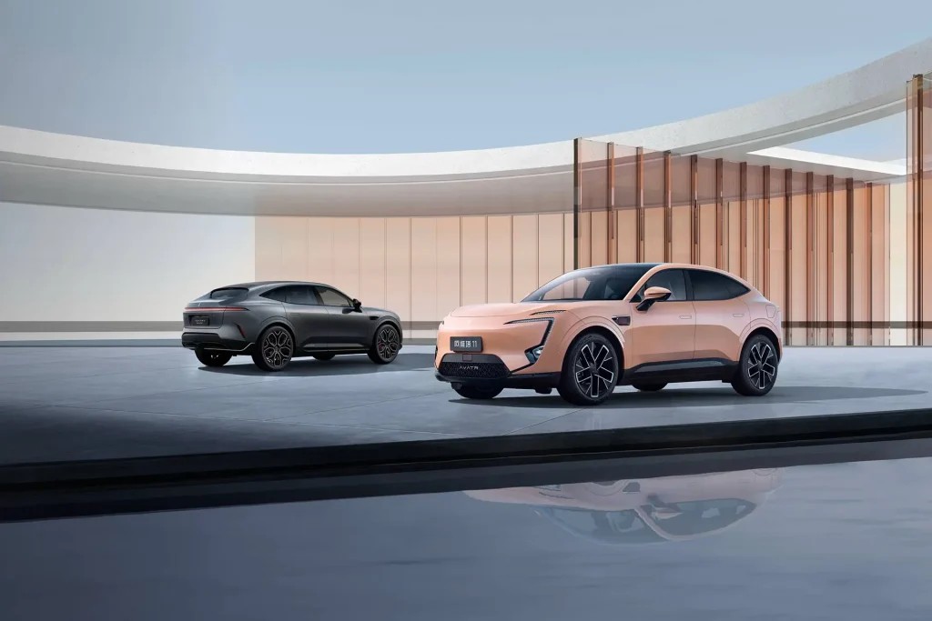 Dois carros esportivos estacionados em uma plataforma com acabamento moderno: um modelo branco na frente e um cinza atrás, em ambiente arquitetônico com estrutura de madeira e vidro ao fundo.