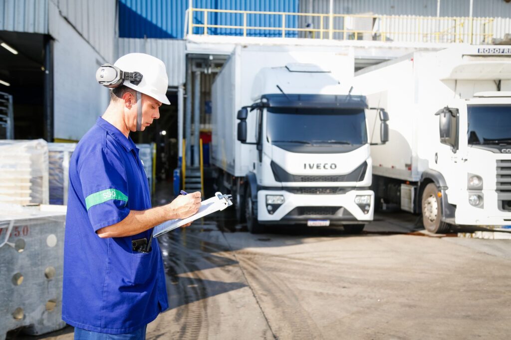 Homem em ambiente industrial com capacete de segurança e uniforme azul, verificando e anotando informações em uma prancheta ao lado de caminhões Iveco estacionados no pátio de logística.