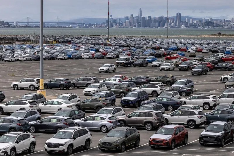 Estacionamento lotado com centenas de carros alinhados em um pátio, vistos de cima, com linha do horizonte da cidade ao fundo.