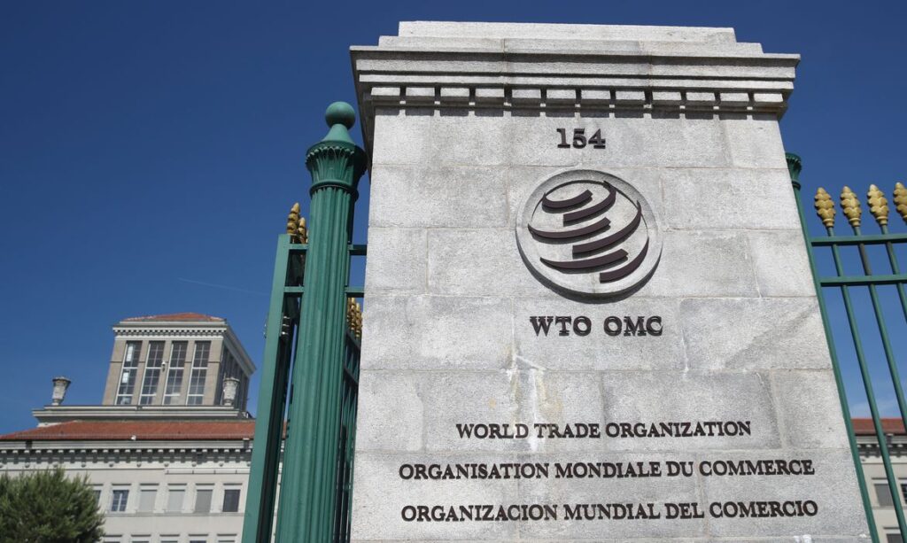 Fachada de prédio com o logotipo e a inscrição da WTO OMC (World Trade Organization), com número 154, em fachada de instituição em cenário externo ao céu azul.
