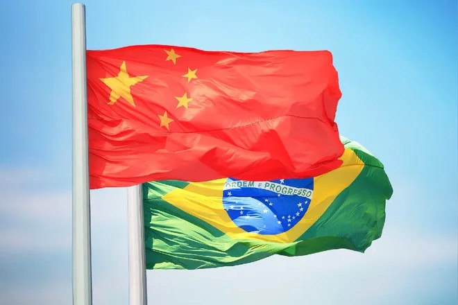 Bandeiras do Brasil e da China tremulando ao vento em um céu azul, com o fundo desfocado e as cores vermelho, amarelo e verde em destaque