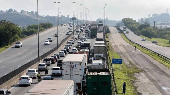 Vista aérea de uma rodovia movimentada com carros e caminhões em fila, faixas múltiplas e barreiras laterais, com caminhões ao centro e tráfego intenso ao fundo.