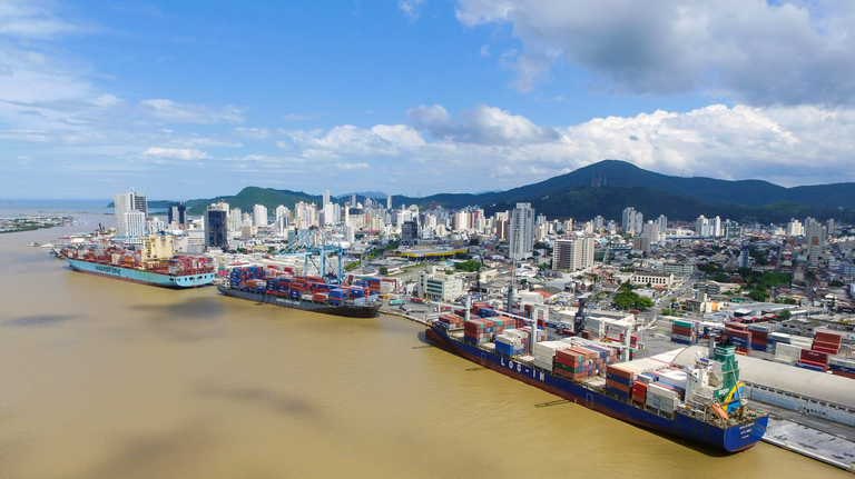 Vista aérea de um porto com navios cargueiros atracados, contêineres empilhados e cidades ao fundo, com montanhas e céu parcialmente nublado no litoral.