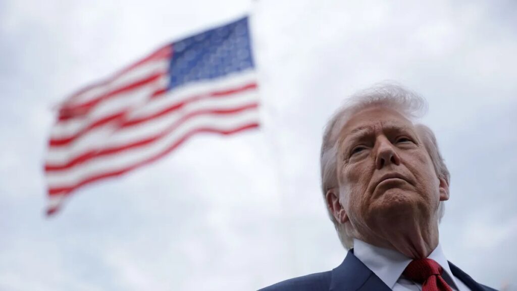 Donald Trump em traje social observa ao lado de uma bandeira dos Estados Unidos ao fundo, em uma imagem ao ar livre com céu nublado e clima político.