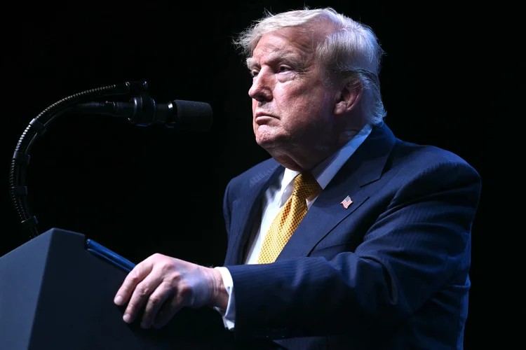 Donald Trump aparece em um palco, sentado e olhando para o lado, durante uma entrevista ou discurso, com um microfone à esquerda e iluminação escura ao fundo.