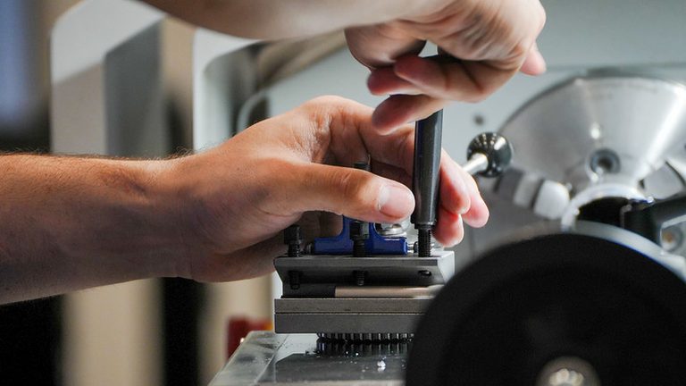 Mãos ajustando uma peça em uma máquina industrial, com os dedos segurando e fixando um componente durante o processo de montagem em oficina