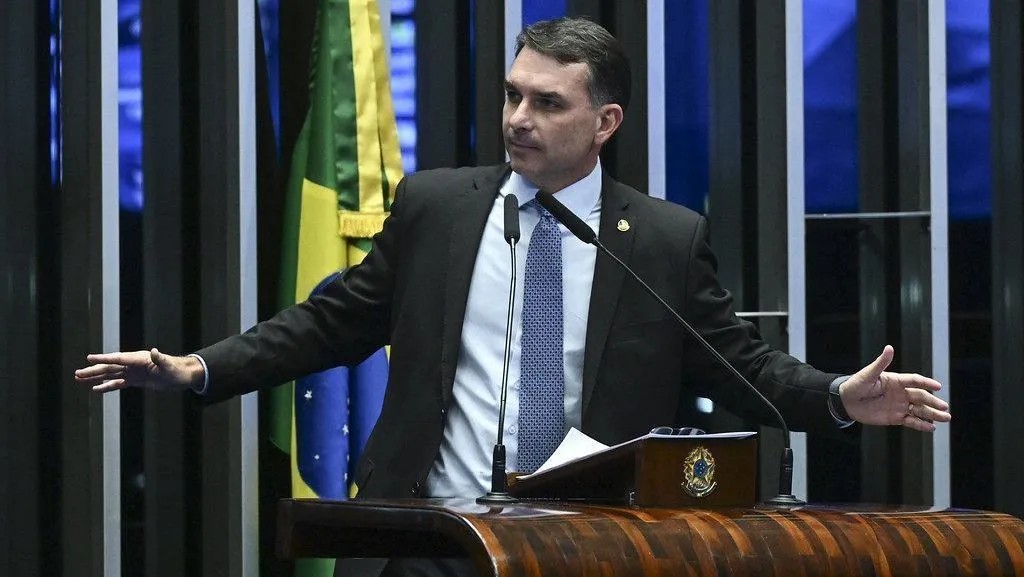 Homem em traje social preto discursa em plenário, com microfone e mesa de madeira, braços abertos ao lado de bandeira do Brasil e painel ao fundo.