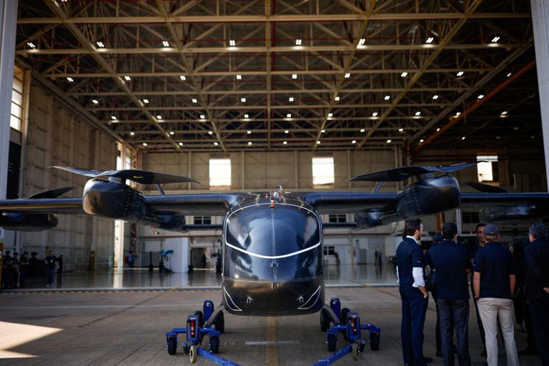Vista frontal de um avião de combate estacionado em um hangar, com o trem de pouso e hélices visíveis, enquanto pessoas observam dentro do galpão industrial.