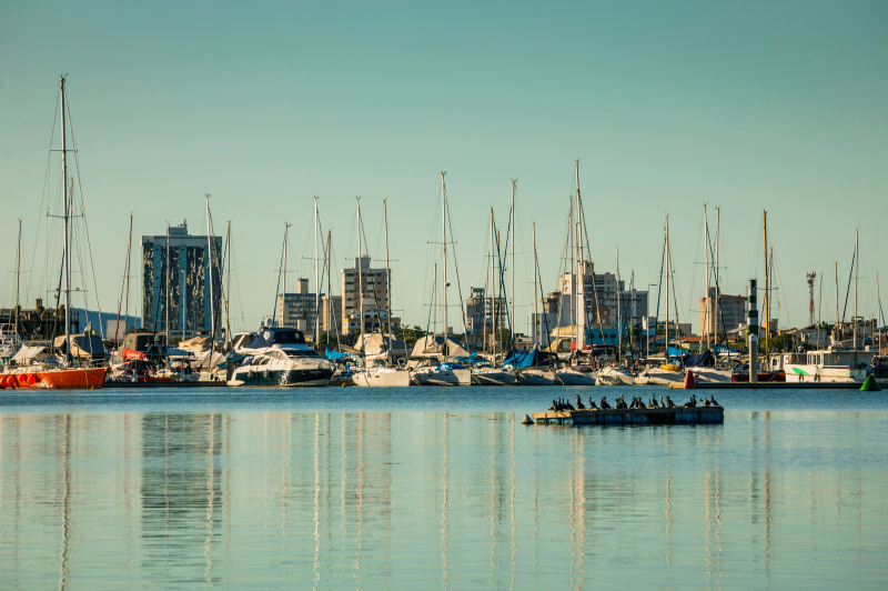 Vista de marina com muitos barcos e iates ancorados, ao lado de uma faixa de cais e prédios na cidade ao fundo, sob céu claro