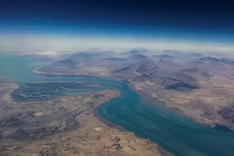 Vista aérea de um grande rio azul serpenteando pelo terreno, com montanhas e áreas desérticas ao fundo, capturada ao amanhecer ou fim de tarde.