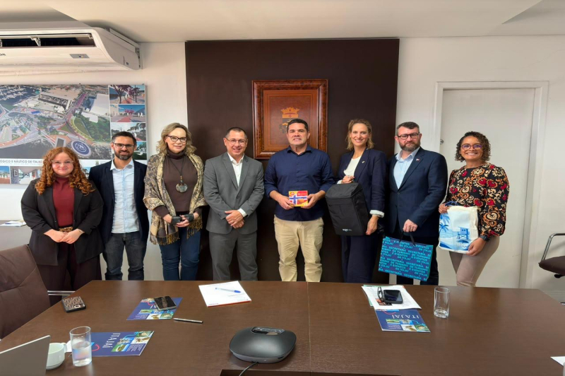 Foto de grupo em ambiente corporativo, com sete pessoas em frente a uma mesa e mural ao fundo, durante reunião oficial com documentos e brindes.