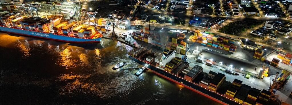 Vista aérea noturna de um porto comercial com contêineres coloridos, navios atracados e iluminação ao longo do cais, mostrando o fluxo de carga e logística.