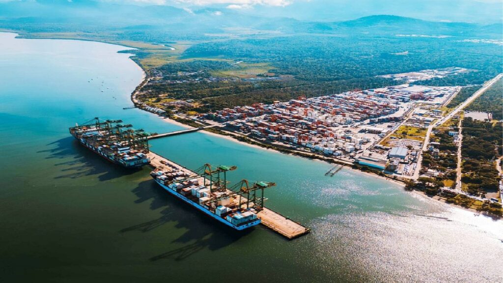 Vista aérea de um porto marítimo com contêineres empilhados em navios e instalações na costa, com rio/baía e cidade ao fundo