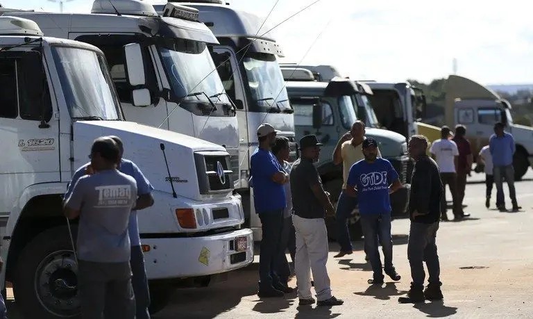 Pessoas em frente a uma fila de caminhões e vans brancas em área externa, com os veículos alinhados lado a lado e trabalhadores aguardando ao sol.