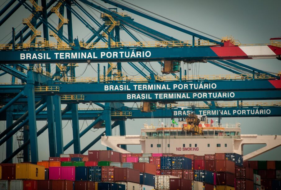 Porto com guindastes e navio de carga ao lado de contêineres, com placas “BRASIL TERMINAL PORTUÁRIO” na estrutura metálica.