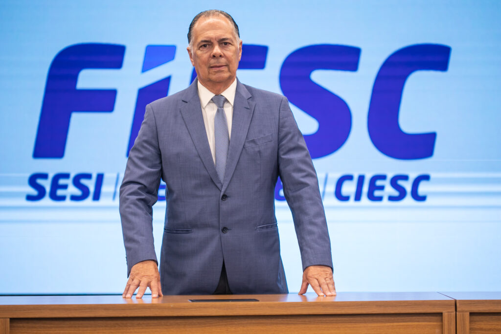 Imagem de um homem em traje social, em pé atrás de um púlpito, com expressão séria, diante de um telão azul com as siglas FIESC e SESI e a palavra CIESC.