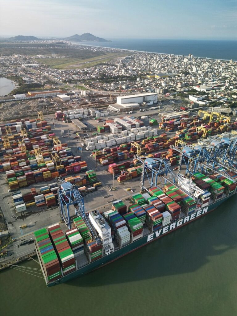 Vista aérea de um navio porta-contêineres no porto, com pilhas de contêineres coloridos e o texto “EVERGREEN” no casco, ao lado de áreas urbanas e mar ao fundo.