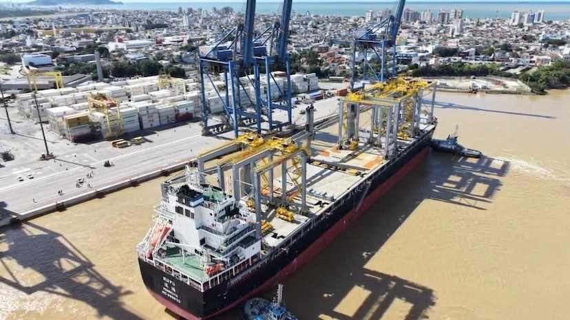Vista aérea de um navio cargueiro no porto, com guindastes e estruturas industriais ao lado do cais, tanque de amarração e movimentação em áreas de armazenamento.