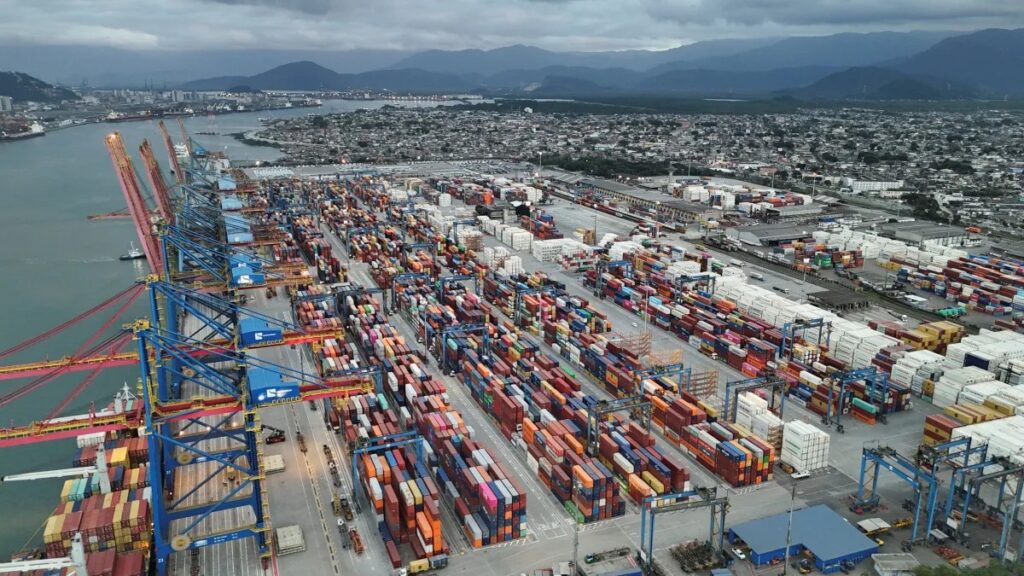 Vista aérea de um grande porto marítimo com contêineres coloridos empilhados em pátios e navios atracados ao lado, mostrando a movimentação logística e a infraestrutura portuária.