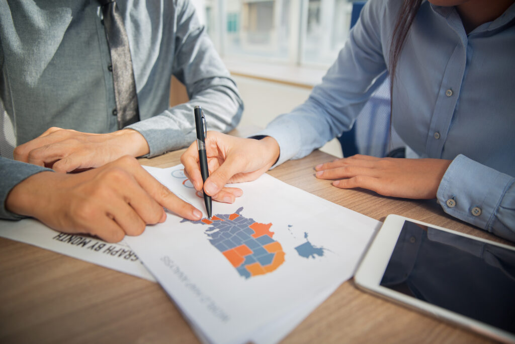 Equipe analisando um mapa e usando canetas para discutir dados em reunião, com gráficos e regiões coloridas em uma folha sobre a mesa e tablet ao lado.