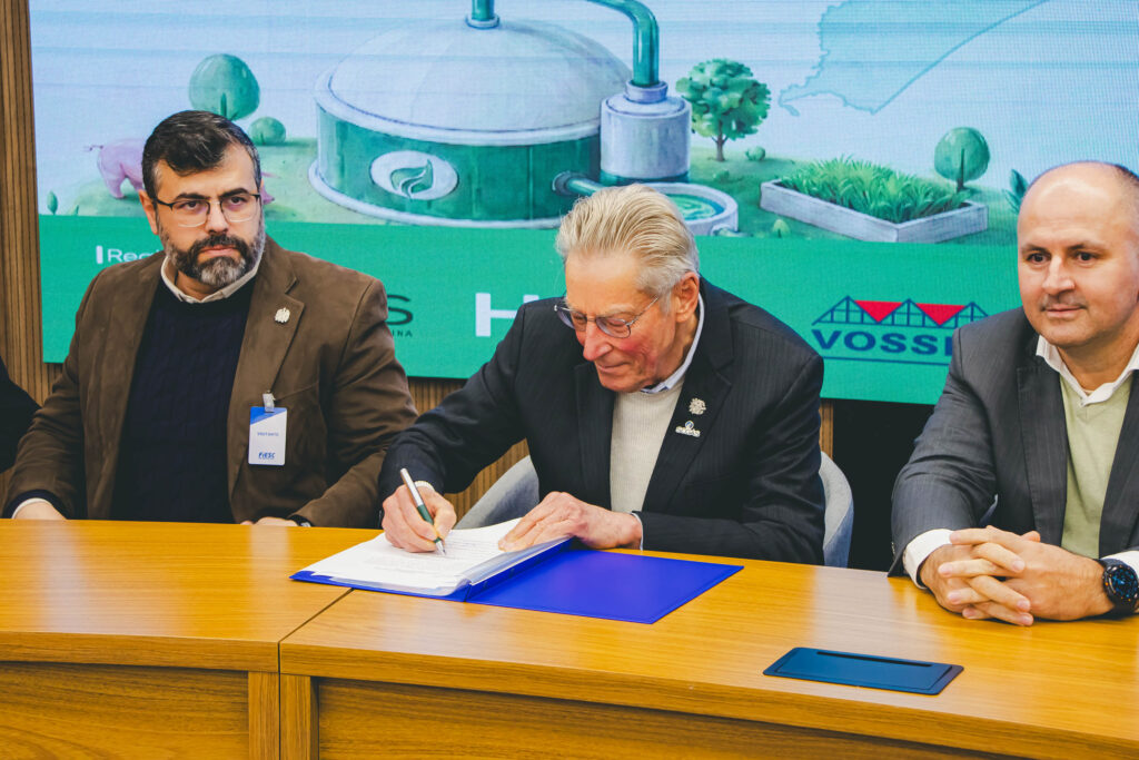 Três homens sentados à mesa durante assinatura de documento em evento corporativo, com fundo de painel verde e tecnologia relacionada a energia, água e agricultura.