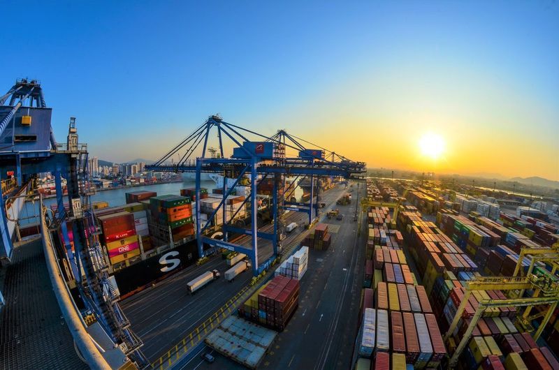 Vista aérea de um grande porto de contêineres no pôr do sol, com navio porta-contêineres ao lado de pilhas de carga e guindastes movimentando e organizando as remessas