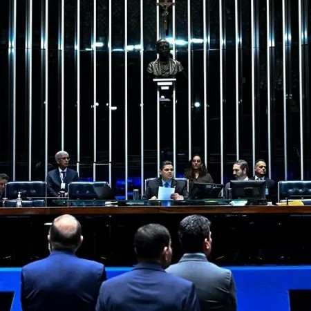 Sessão do plenário de um tribunal com autoridades e advogados sentados, vista do público em frente ao corpo legislativo e uma estátua ao fundo entre colunas iluminadas.