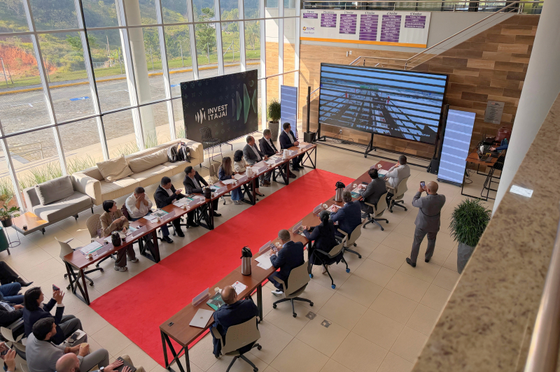 Sala de reunião em evento corporativo com mesas em formato retangular, cadeiras e um telão, participantes sentados e pé-direito alto com janelas amplas ao fundo.