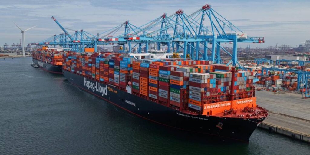 Navio porta-contêineres Hapag-Lloyd atracado em terminal portuário, com pilhas de contêineres coloridos e guindastes ao fundo, em vista aérea sobre o cais.