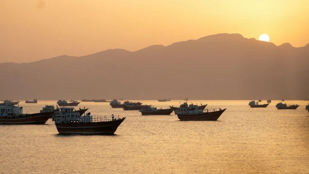 Pôr do sol sobre um mar calmo com vários barcos e navios ancorados, ao fundo montanhas e atmosfera dourada na paisagem costeira.