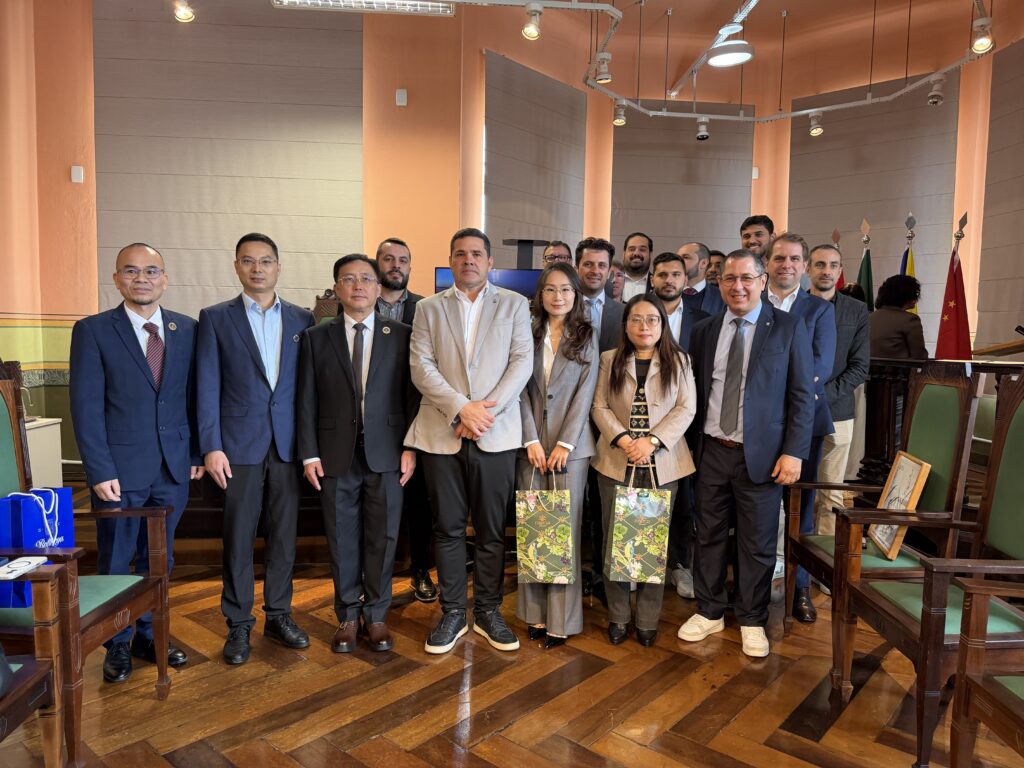 Foto de grupo de políticos e autoridades em um ambiente institucional, com vários homens e mulheres em trajes sociais, sentados e em pé, diante de mesa e bandeiras ao fundo, em auditório.