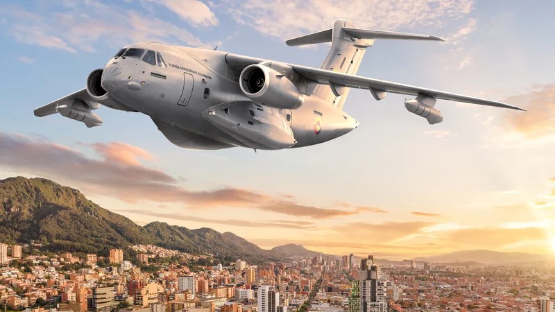 Aeronave de transporte militar decolando sobre uma cidade ao entardecer, com montanhas ao fundo e céu com nuvens, mostrando jatos e motores em destaque.