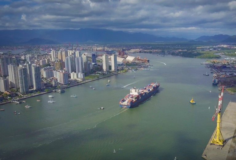 Vista aérea de um porto e canal de navegação com navios e contêineres, com prédios na margem e montanhas ao fundo sob céu nublado.
