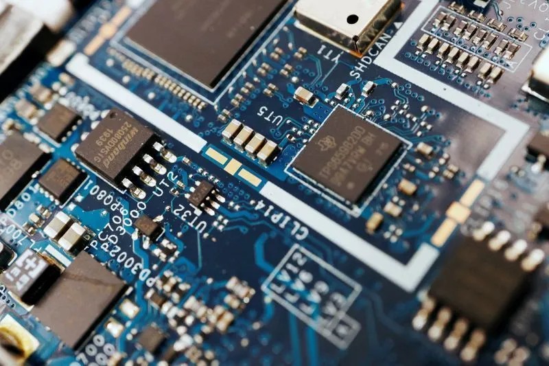 Close-up de uma placa de circuito eletrônica com componentes SMD, trilhas e chips integrados azul e branco, mostrando detalhes de soldas e conectores.