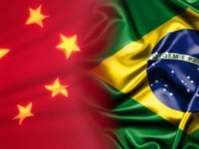 Bandeiras do Brasil e da China tremulando ao vento, com a bandeira de fundo verde e amarela e a bandeira com estrelas vermelhas ao lado, em imagem desfocada.