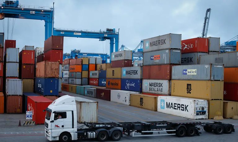 Pátio de contêineres em terminal portuário com caixas de carga empilhadas e caminhão transportando mercadorias, com contêineres da Maersk
