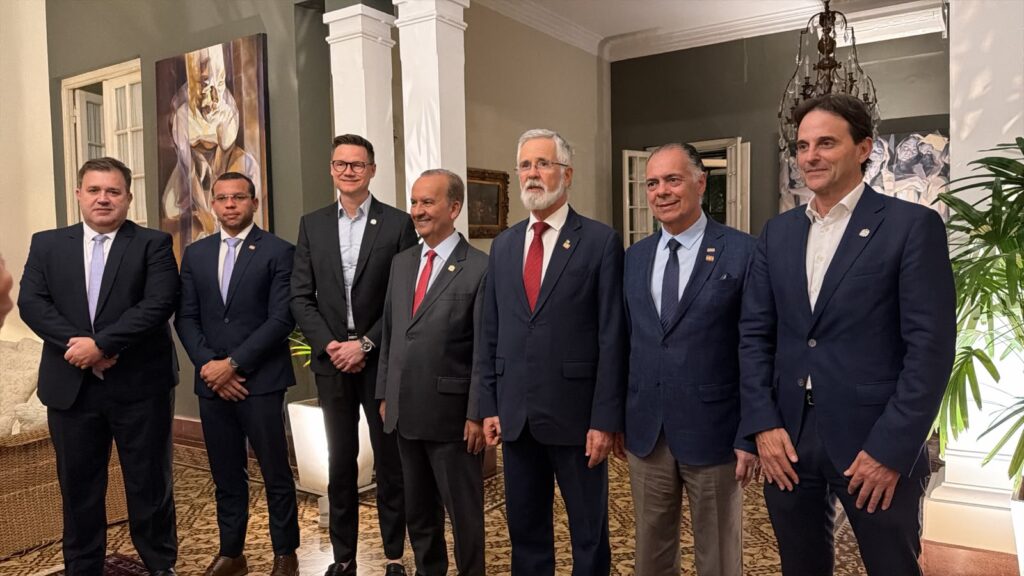 Grupo de autoridades em trajes formais, posando para foto em um ambiente interno com colunas brancas e decoração elegante ao fundo.