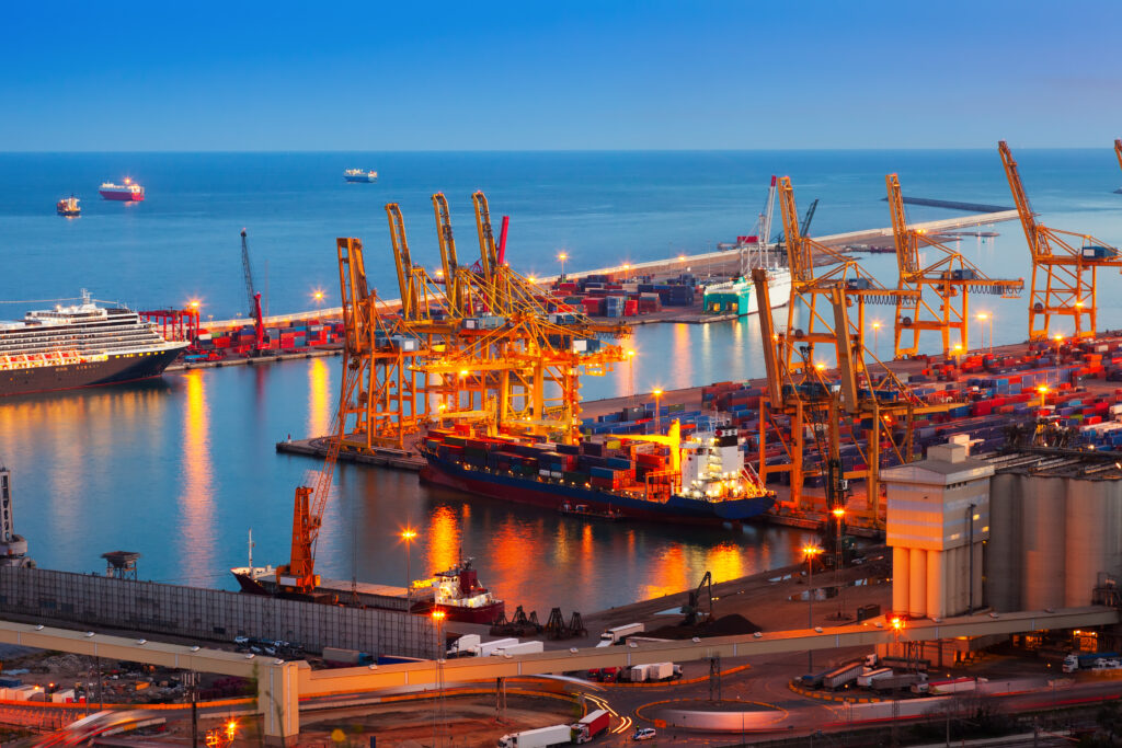 Vista do porto marítimo ao entardecer, com guindastes laranjas, áreas de carga e navios ao lado d’água azul, com iluminação acesa e céu claro.