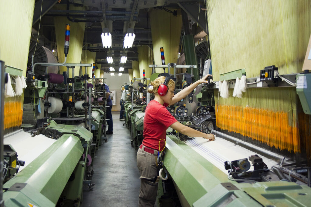 Funcionária em uma fábrica têxtil opera máquinas industriais e ajusta o tear, enquanto fios coloridos passam pelos equipamentos em uma linha de produção.