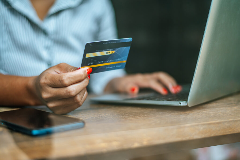 Pessoa usando um cartão bancário na mão enquanto realiza uma compra no computador, em ambiente de trabalho, com smartphone sobre a mesa ao fundo desfocado.