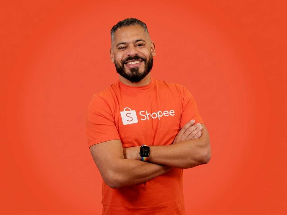 Homem sorridente usando camiseta laranja com o logo da Shopee, braços cruzados, em fundo laranja vibrante. Foto promocional de marca.