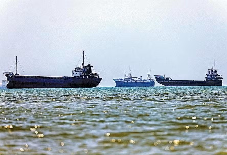 Três navios-tanque e embarcações militares no mar, vistos à distância com o céu nublado e o reflexo das ondas na água, em cenário portuário.